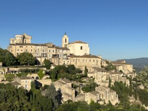 Panoramic view of Gordes hilltop village with golden limestone buildings, cypress trees, terraced gardens, and a château tower under a clear blue sky, Luberon, Provence, France
