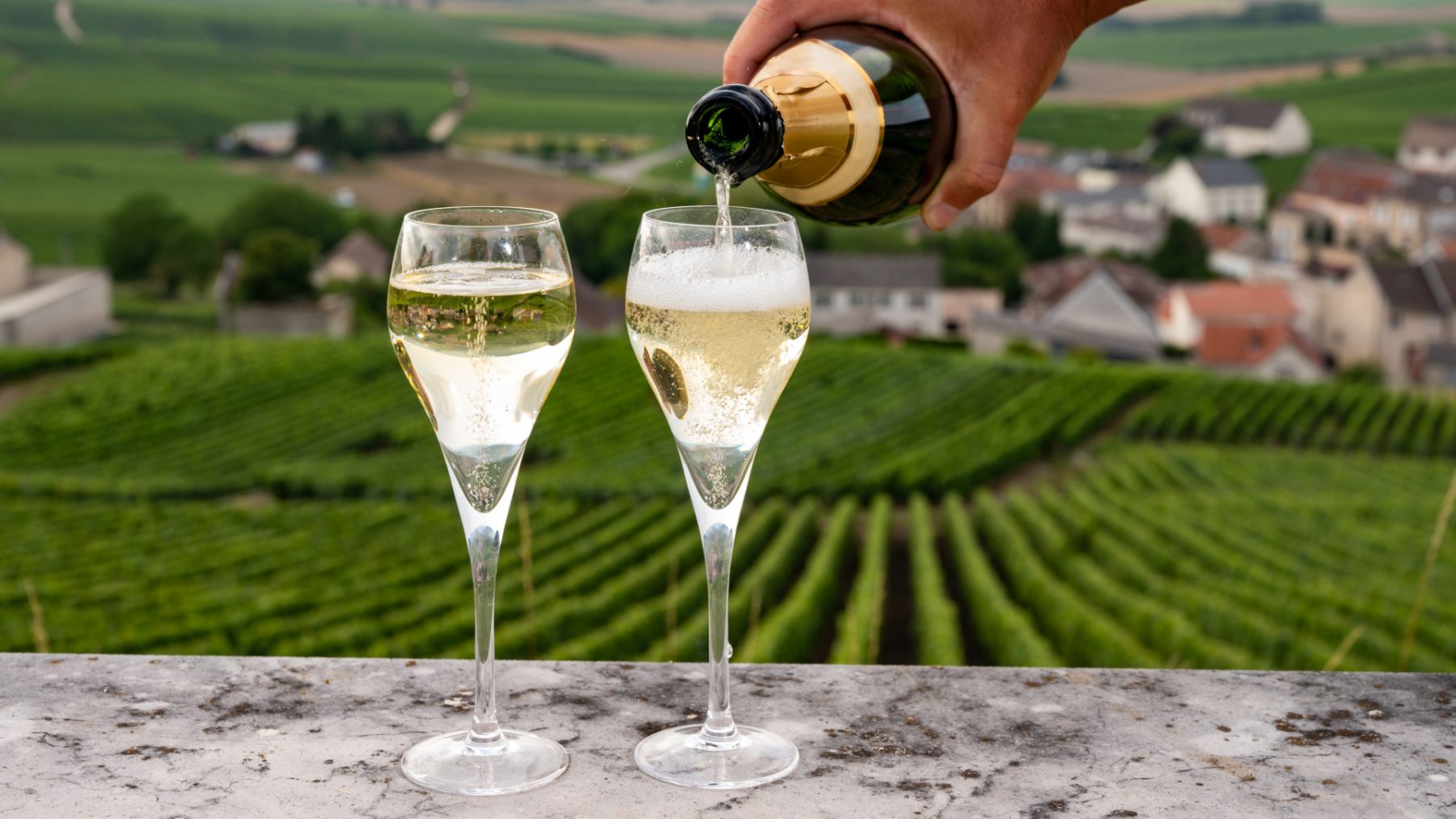 Pouring champagne into two flutes overlooking rolling Champagne vineyards and village on a luxury Champagne wine tour, France