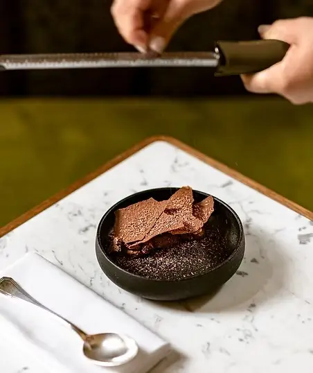 Chef grating dark chocolate over a chocolate mousse dessert with shards in a black ceramic bowl on a white marble board, fine dining at a French château