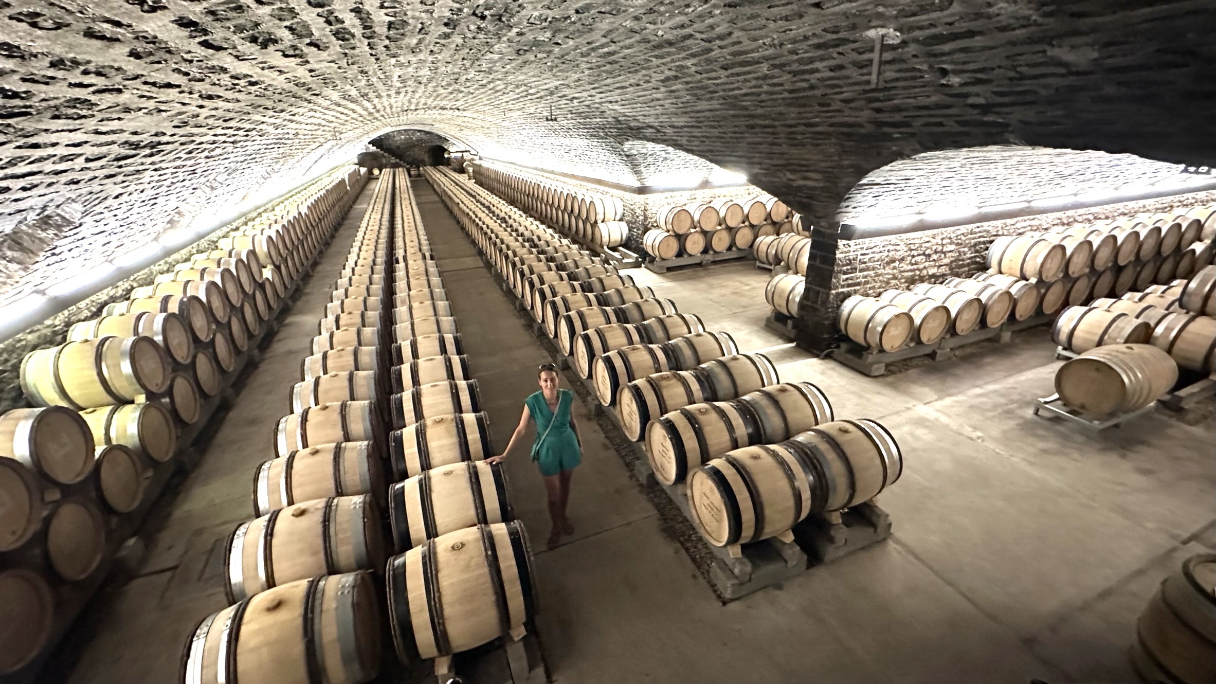 Stephanie Burtin walking through vast Burgundy Grand Cru cellar with hundreds of oak barrels in vaulted stone tunnels on a luxury wine tour, France