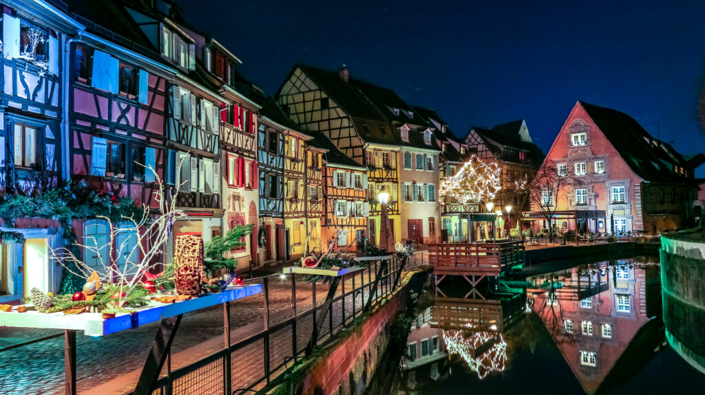 Colmar Christmas market canal at night with colourful illuminated half-timbered houses and festive decorations reflected in water, Alsace, France