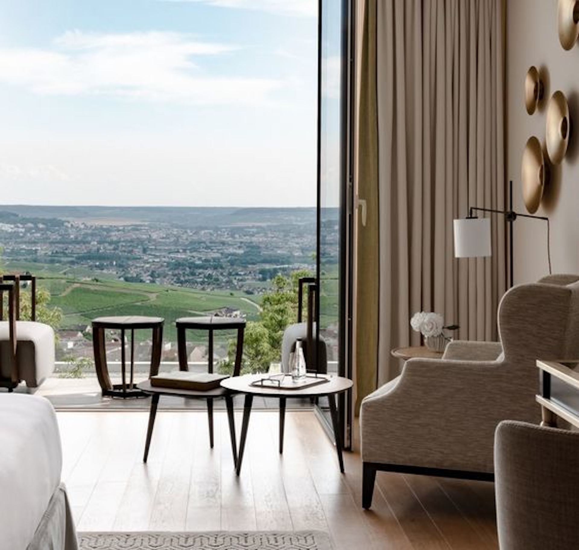 Upscale hotel suite with floor-to-ceiling windows overlooking rolling vineyard hillsides, featuring neutral linen armchairs, dark wood accent tables, warm hardwood flooring, and brushed gold wall art